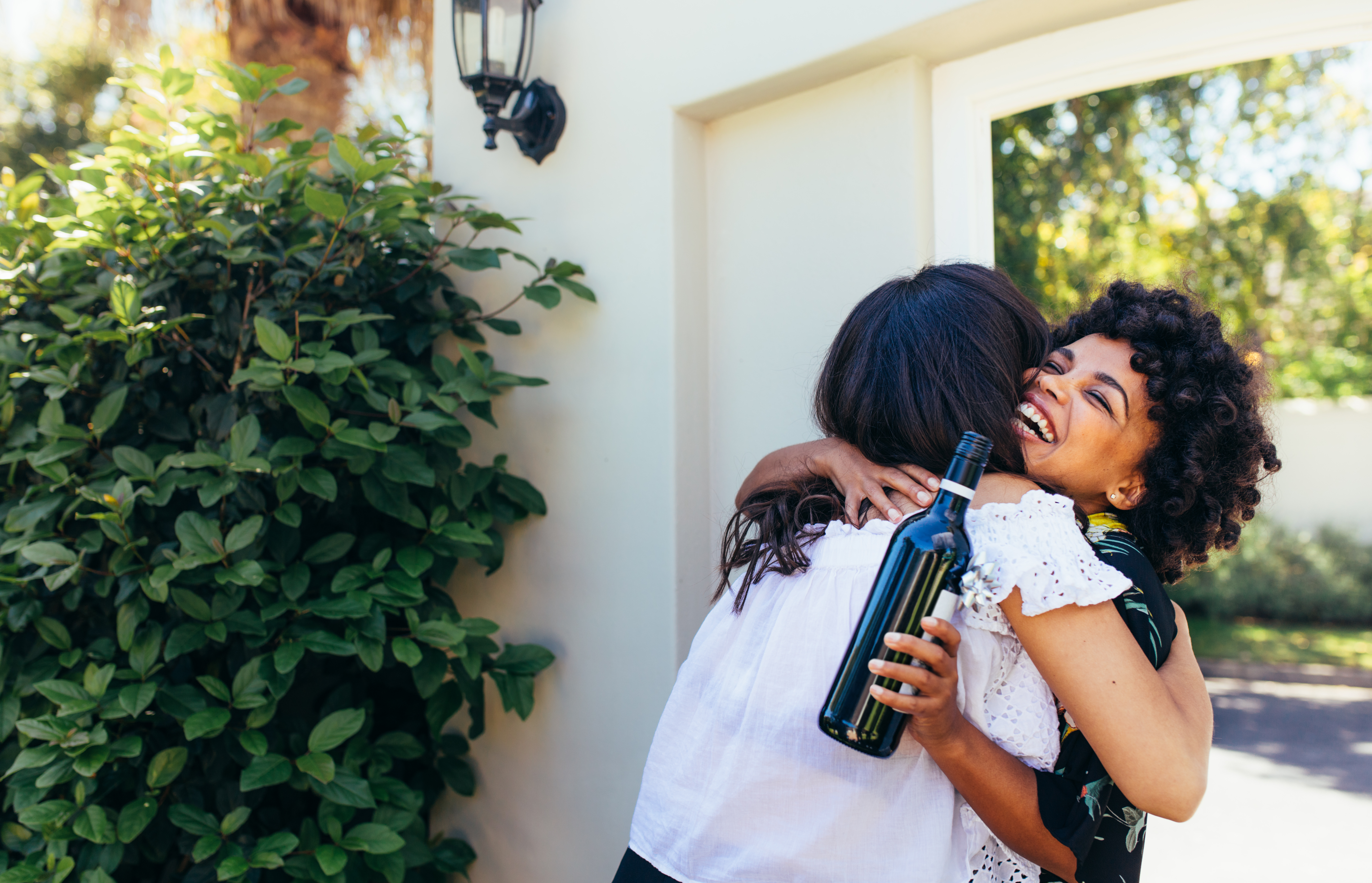 African woman greeting and embracing female friend for having a new house. Smiling young woman with wine bottle congratulating her friend. Housewarming with friends.