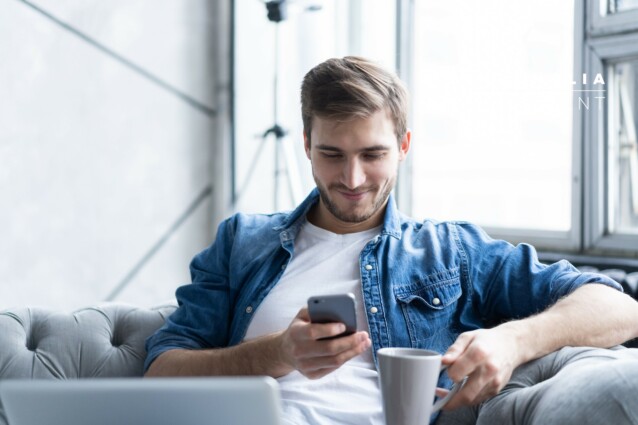 Young man using his smartphone for online banking - sitting on sofa with laptop on leap